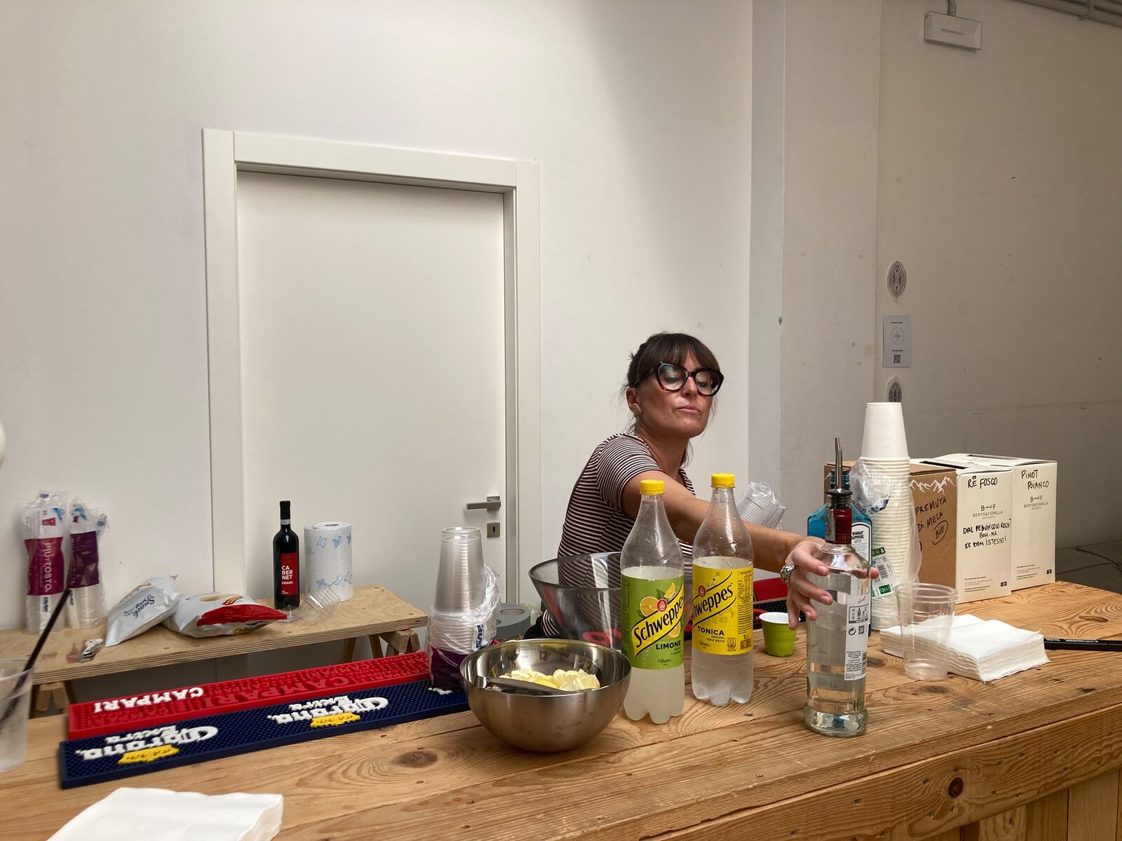 Laura G. dietro il bancone di Zephiro mentre prepara il rinfresco: tappetini Campari, bottiglie di Schweppes e tonica, prosecco, una ciotola di patatine, bicchieri di carta in pila, sguardo concentrato. Dieci secondi rubati al lavoro del bar, da una che il bar lo ha nel sangue