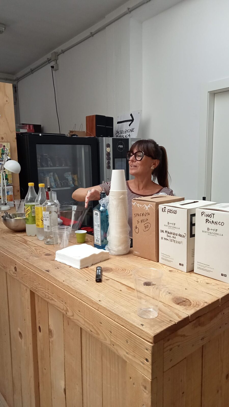 Laura G. dietro il bancone di Spazio Zephiro alle 14:47, mentre prepara il rinfresco prima dell&rsquo;arrivo: bottiglie di prosecco e gin sui ripiani, cartoni di Pinot Bianco e refosco, pile di bicchieri di carta, una bottiglia di spremuta di mela. Lei al centro, occhiali, capelli raccolti, gesto a metà — il bar che si arma